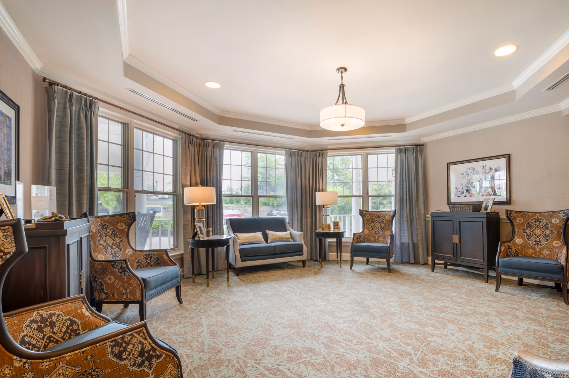 Bright seating room with patterned armchairs, a small sofa, side tables and large windows.