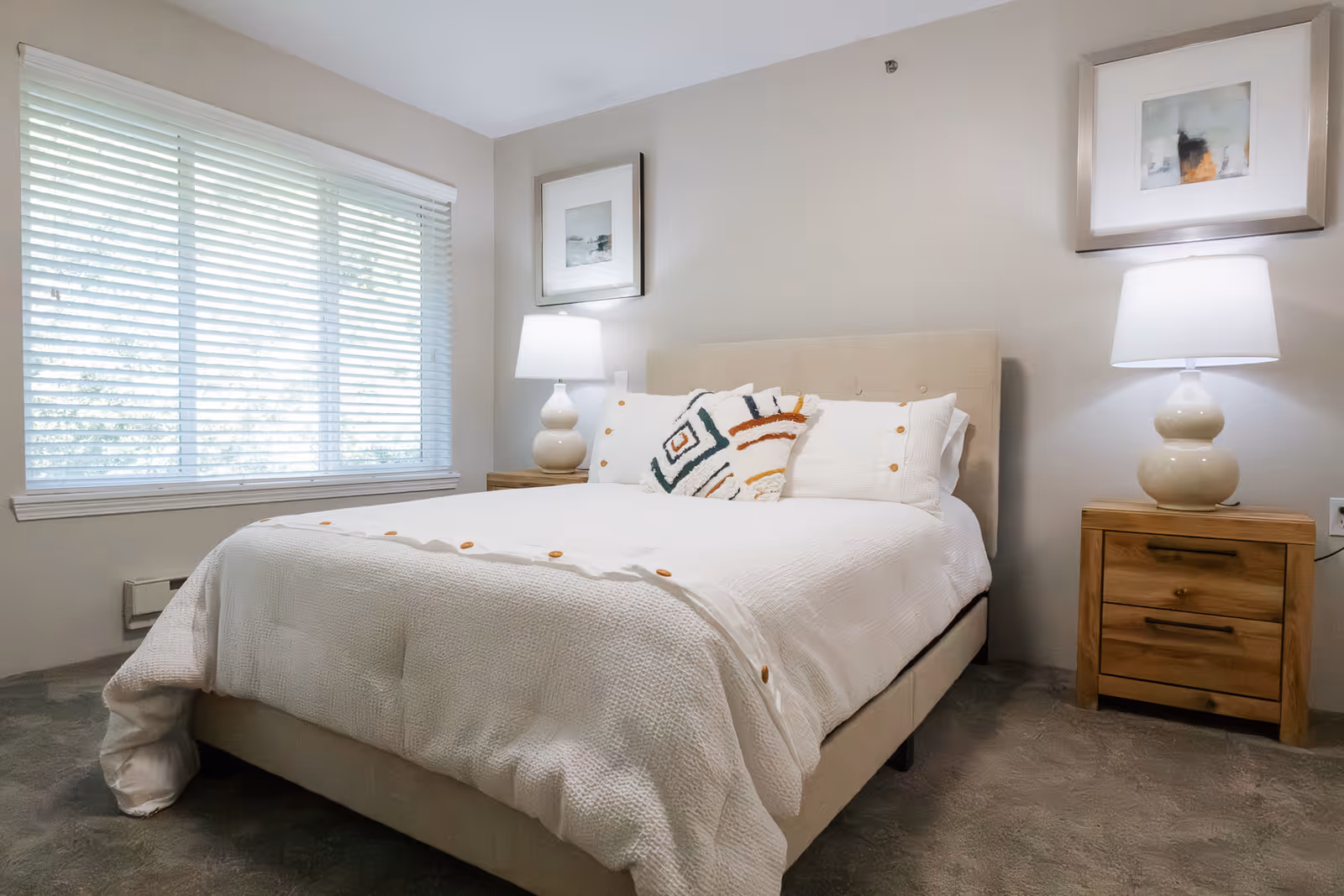 A neatly made bed with white bedding and decorative pillows in a bedroom. There are two wooden nightstands on either side of the bed, each with a beige lamp. Two framed pictures hang on the wall above the nightstands. A window with white blinds is on the left side of the room, allowing natural light to enter.