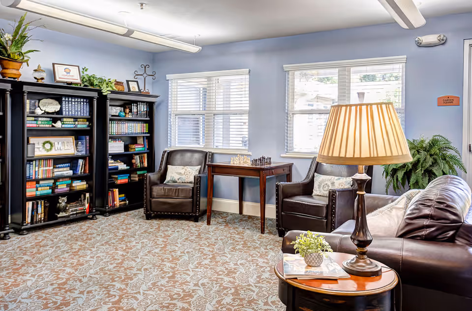 A cozy living room area with two dark leather armchairs and a matching leather sofa. Between the armchairs is a wooden table with a chess set on it. There are two large windows with white blinds letting in natural light. A wooden side table with a lamp and a small plant is next to the sofa. Two black bookshelves filled with books and decorative items are against a light blue wall. The floor is covered with a patterned carpet, and there are some plants and framed pictures on top of the bookshelves.