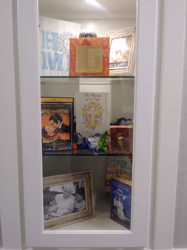 A white display cabinet with glass shelves containing various decorative items including framed photos, books, a DVD of Gone with the Wind, a floral card with a cross, and other small decorative pieces.