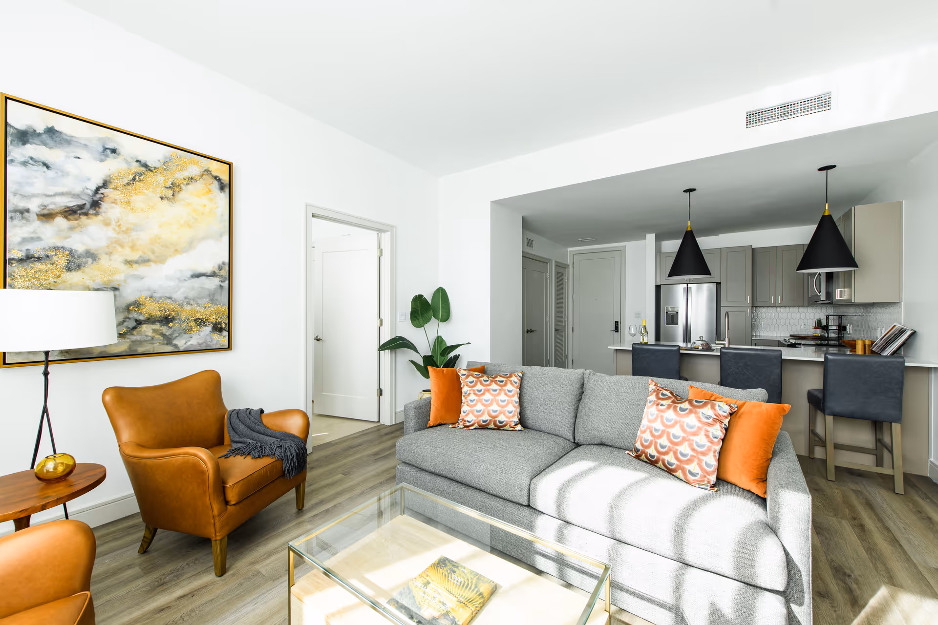 Bright and modern living room with a gray sofa adorned with orange and patterned pillows, a brown leather armchair with a dark throw, a glass coffee table, a side table with a lamp, and a large abstract painting on the wall. The room opens into a kitchen area with gray cabinets, stainless steel appliances, and three black pendant lights hanging over a breakfast bar with three dark chairs.