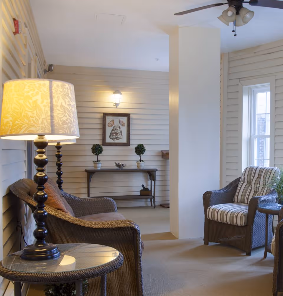 A cozy indoor seating area with wicker furniture including a sofa and armchair with cushions, a glass-top side table with a decorative lamp, a ceiling fan, and a console table against a wall with two small potted plants and a framed artwork. The walls have horizontal paneling and there is a window letting in natural light.
