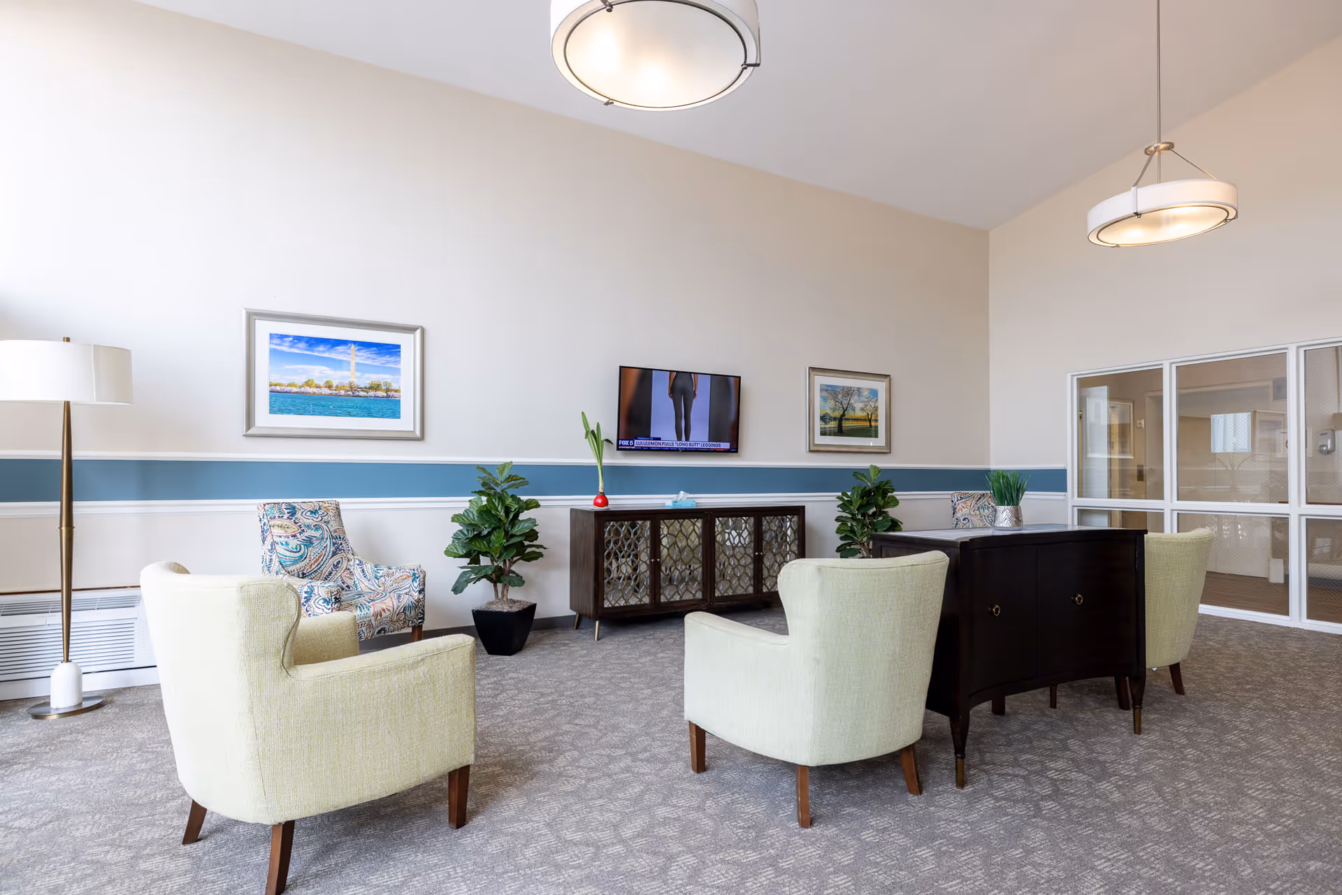 A bright and spacious living room area with light-colored walls and carpeted floor. The room features two light green armchairs, a patterned armchair, a dark wooden console table with decorative plants, a flat-screen TV mounted on the wall, two framed pictures, a floor lamp, and large windows with glass panels on one side.