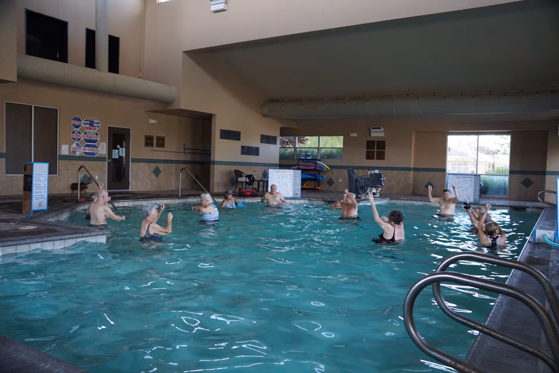 A group of elderly people participating in a water exercise class in an indoor swimming pool. The pool area has large windows, tiled walls, and handrails leading into the water. The participants are spaced out and appear to be following an instructor.