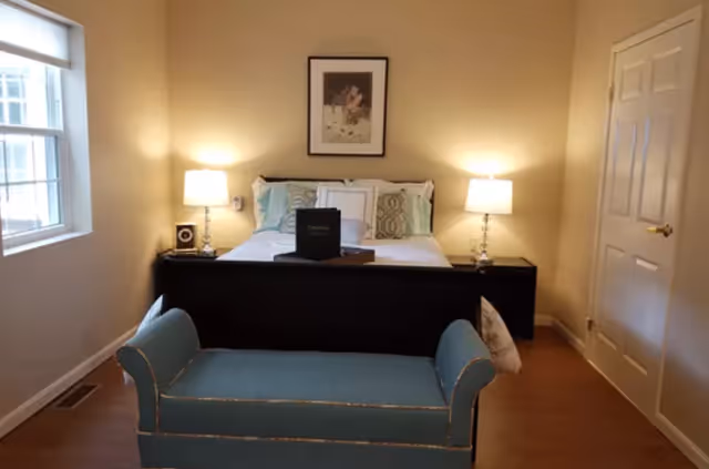 Well-lit bedroom with a bed flanked by two bedside lamps, a teal bench at the foot, and a window on the left.