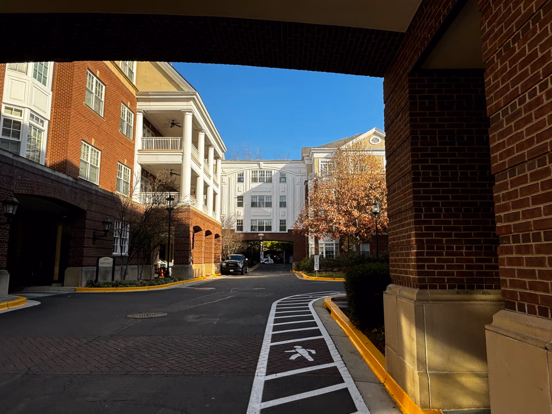 View of the exterior of a senior living facility showing brick buildings with white balconies and columns, a driveway with pedestrian markings, and a tree with autumn leaves under a clear blue sky.