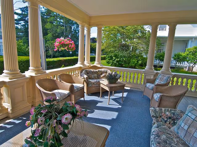 A covered outdoor porch area with wicker furniture including chairs and a loveseat with floral cushions, a small table, and potted plants. The porch has cream-colored columns and railings, overlooking a green garden with trees and shrubs.