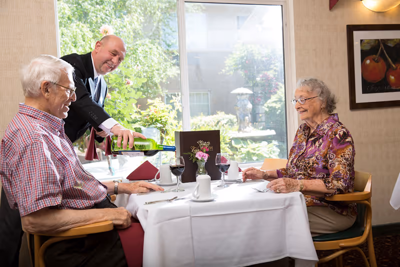 An elderly man and woman sit at a dining table with white tablecloth in a well-lit room with a large window showing greenery outside. A waiter is pouring red wine into the man's glass. The table is set with wine glasses, coffee cups, silverware, and a small flower vase. The woman is smiling and wearing a patterned blouse.