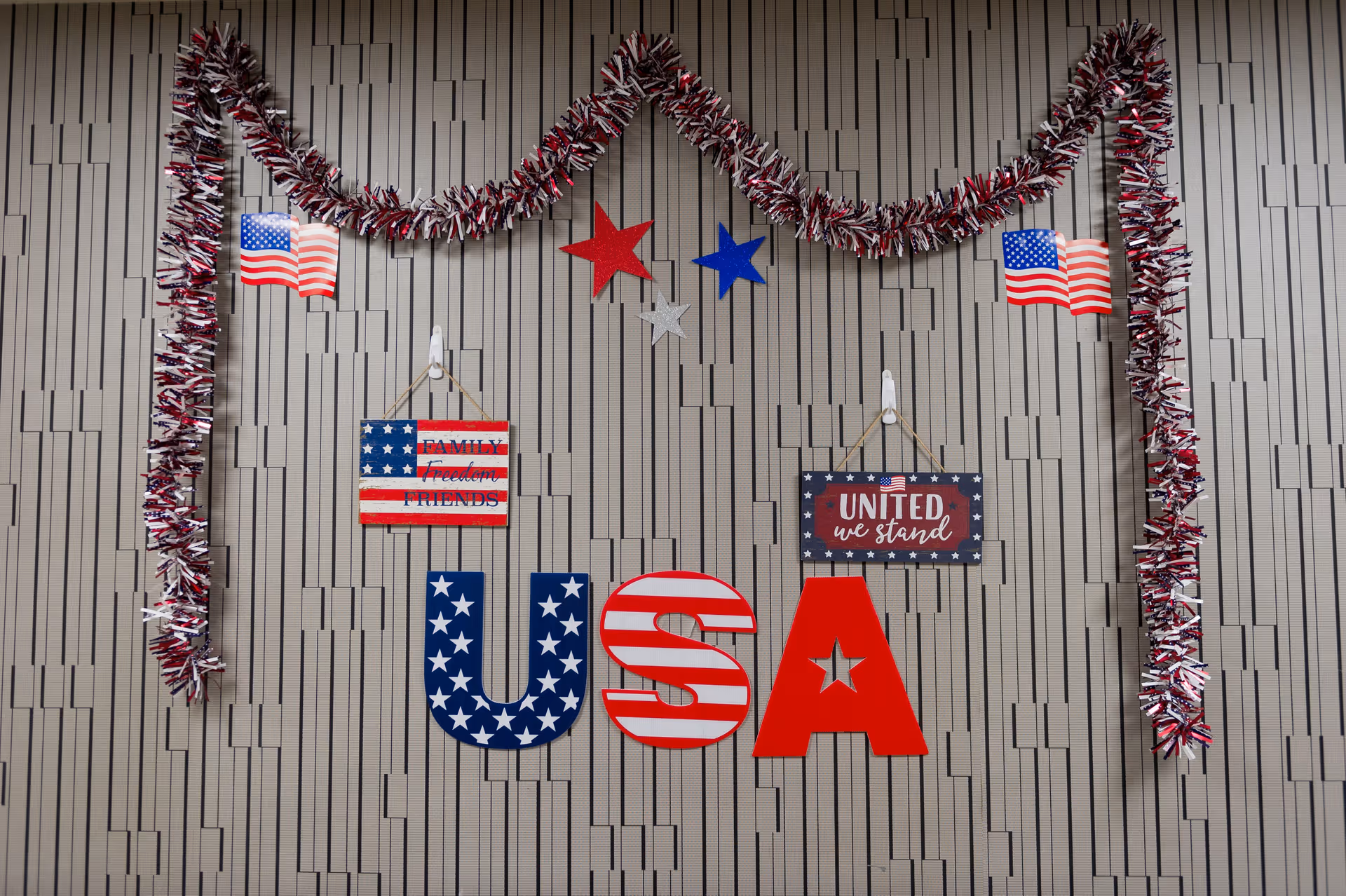 Wall decorated with patriotic USA-themed items including red, white, and blue tinsel garlands, small American flags, red, white, and blue stars, and large letters spelling 'USA' with stars and stripes patterns. Two hanging signs read 'FAMILY Freedom FRIENDS' and 'UNITED we stand'.