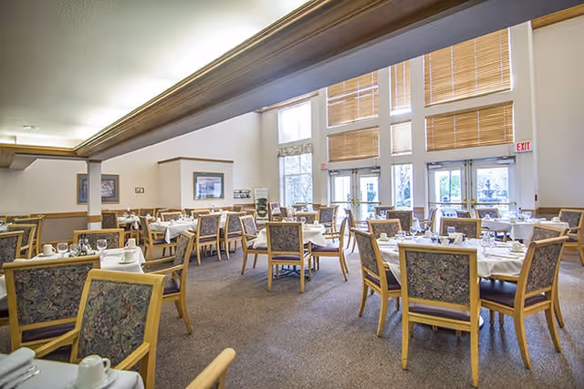 A spacious dining room with multiple tables set with white tablecloths, cups, and saucers. The room has large windows with blinds, allowing natural light to fill the space. Wooden chairs with patterned upholstery surround each table. The walls are decorated with framed artwork, and there is an exit door visible at the far end.