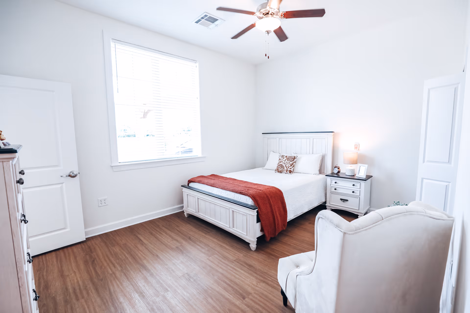A bright and clean bedroom with a white wooden bed frame, white bedding, and a red throw blanket. There is a white nightstand with a lamp, clock, and picture frame next to the bed. A white armchair is positioned in the corner, and a ceiling fan with lights is mounted on the ceiling. The room has wood flooring and a large window letting in natural light.