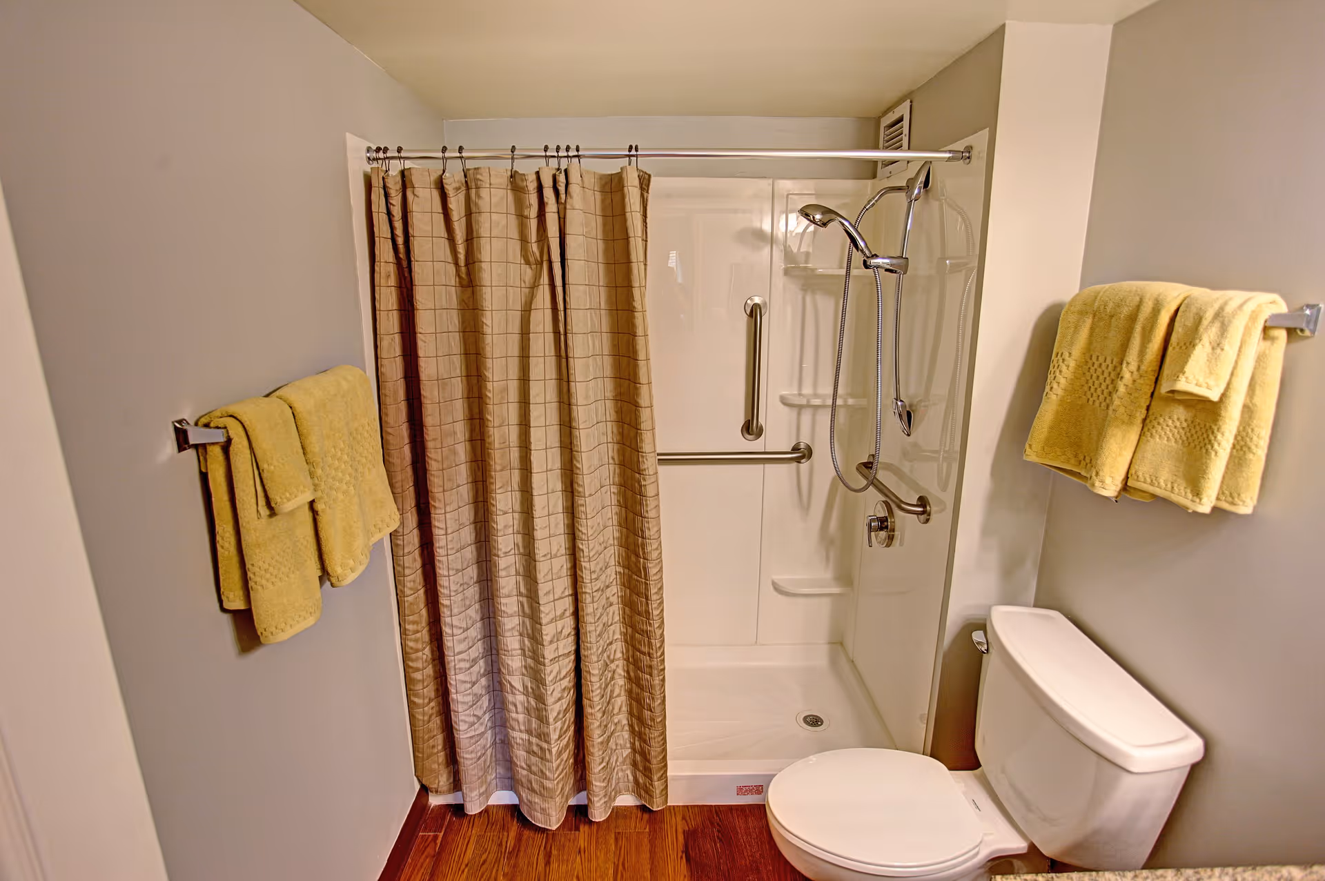 A bathroom with a shower area enclosed by a beige shower curtain. The shower has a handheld showerhead and grab bars for safety. There are yellow towels hanging on towel racks on both sides of the shower. A white toilet is positioned next to the shower, and the floor has a wood-like finish.