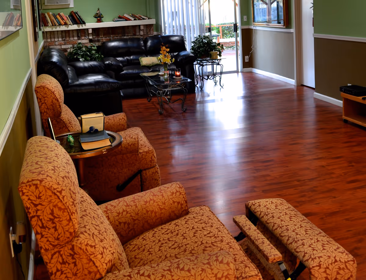 A cozy living room with wooden flooring, featuring two patterned orange recliner chairs in the foreground and a black leather sofa set near a brick fireplace with a white mantle. There are books and decorative items on the mantle, a glass coffee table with flowers and a candle, and a sliding glass door leading to an outdoor patio with greenery visible outside.