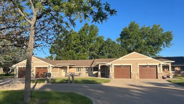 Exterior view of a single-story residential building with beige siding and brown garage doors, surrounded by green trees and a clear blue sky.