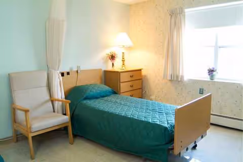 A senior living facility bedroom featuring a single bed, dresser, chair, lamp, and window.