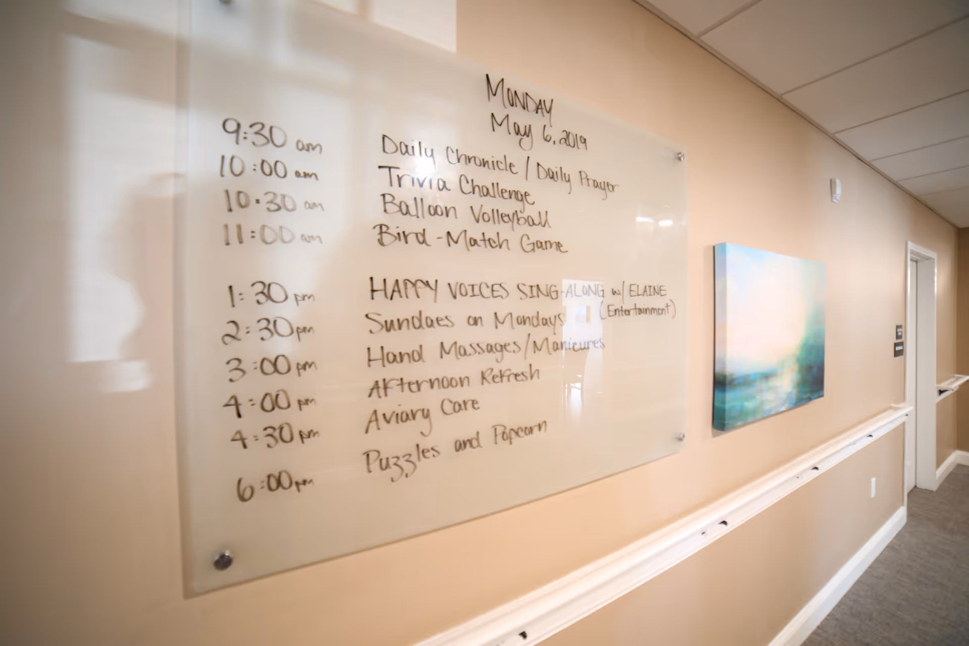 A hallway in a senior living facility with a beige wall and a whiteboard displaying a daily schedule of activities for Monday, May 6, 2019. The schedule includes events such as Daily Chronicle, Trivia Challenge, Balloon Volleyball, Bird-Match Game, Happy Voices Sing Along with Elaine, Sundaes on Mondays, Hand Massages/Manicures, Afternoon Refresh, Aviary Care, and Puzzles and Popcorn. There is also a colorful abstract painting on the wall further down the hallway.
