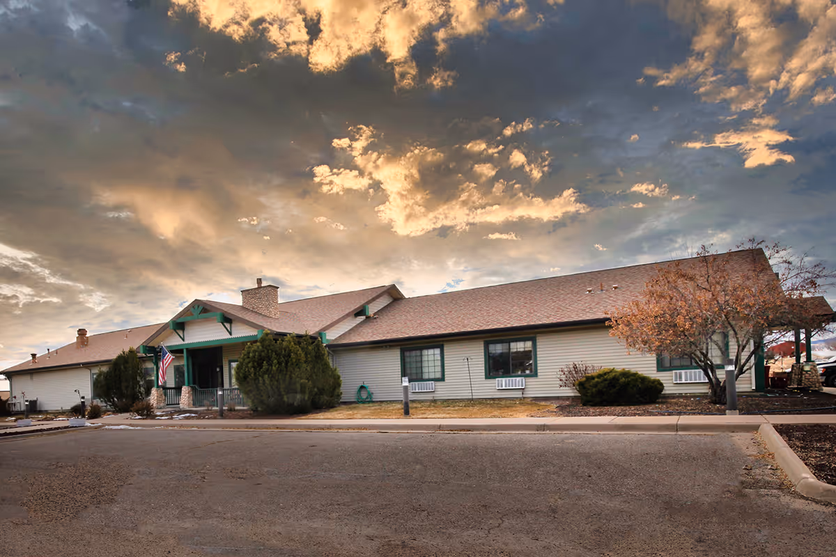 Exterior view of a single-story building with beige siding and a brown roof under a dramatic cloudy sky. The building has green trim around the windows and entrance, an American flag near the entrance, and some trees and bushes in front. The area in front of the building is paved with a parking lot.