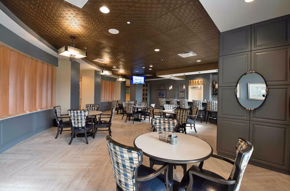 A spacious dining room with multiple round tables and cushioned chairs featuring a blue and white checkered pattern. The room has a decorative brown ceiling, soft lighting fixtures, and large windows with wooden blinds. A wall-mounted TV and a bar area with high chairs are visible in the background.