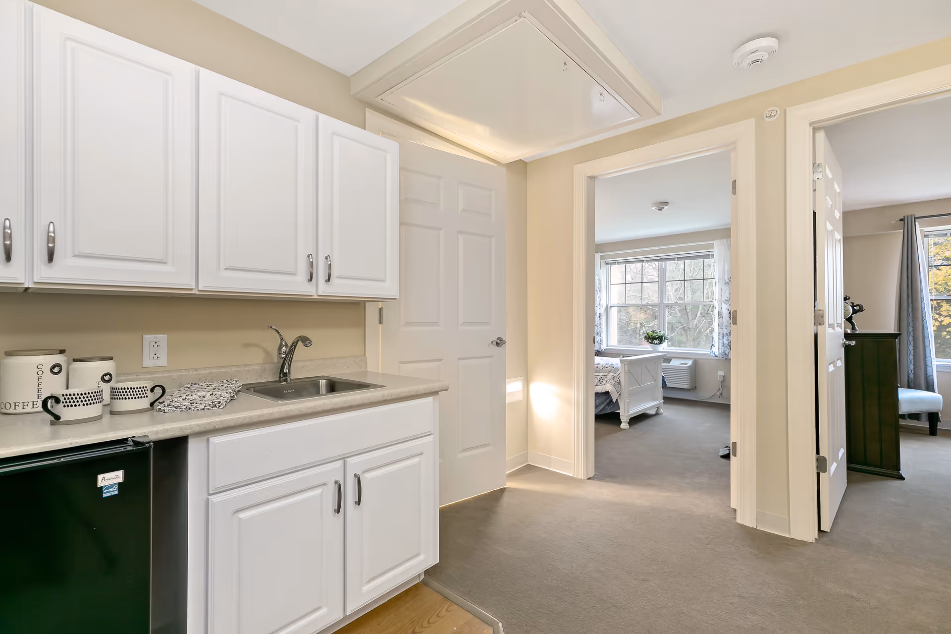 A small kitchenette area with white cabinets, a countertop with a sink, and a mini refrigerator. On the countertop, there are three coffee canisters and a patterned cloth. The kitchenette opens into a hallway with two doorways leading to rooms with large windows letting in natural light.