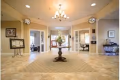 A spacious and well-lit interior common area with beige walls and tiled floors. In the center, there is a round wooden table with a floral arrangement on top, placed on a large patterned rug. The room has multiple doorways leading to other rooms, with framed pictures and decorative items on the walls. A chandelier hangs from the ceiling, providing warm lighting.