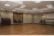 Large multipurpose activity room with wood flooring, chairs along the walls, a piano, bulletin board, and an American flag.