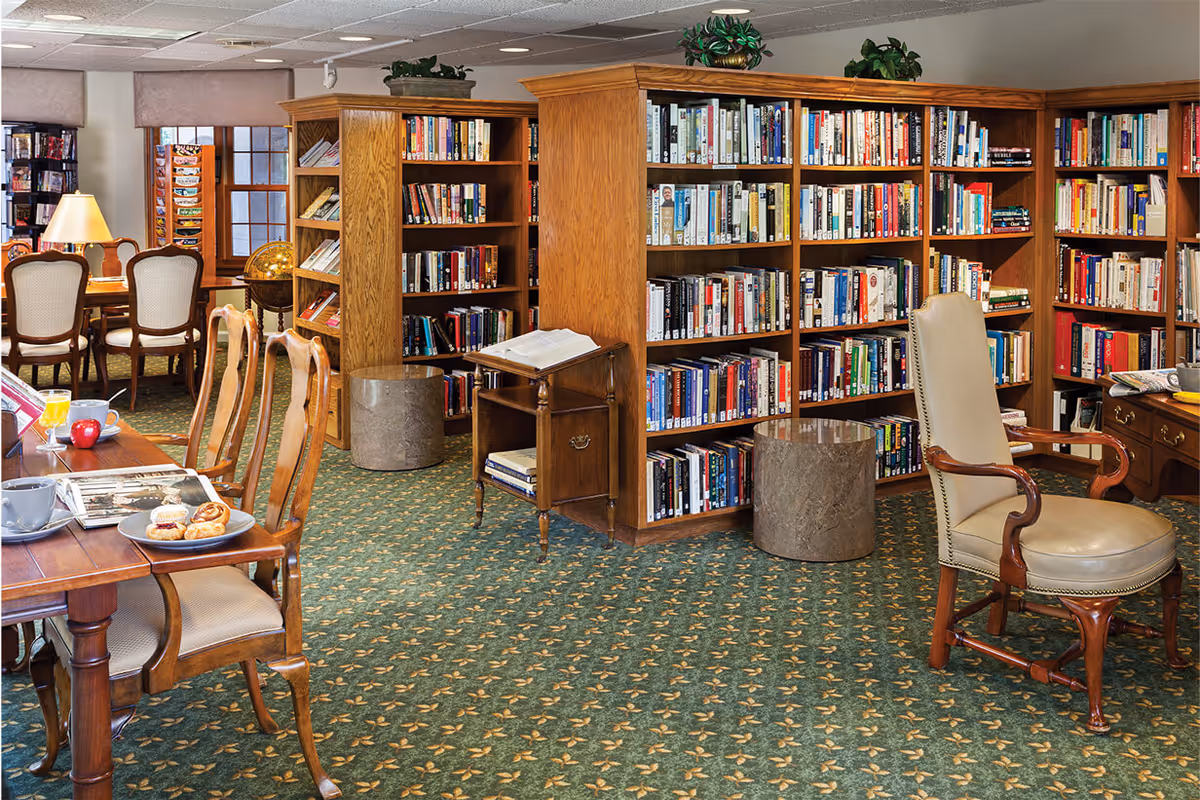Interior view of a library or reading room in a senior living facility with wooden bookshelves filled with books, a green patterned carpet, several wooden chairs, a table with pastries and coffee cups, and a globe near the window.