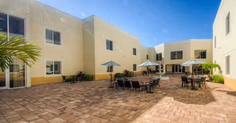 Outdoor courtyard area with paved brick flooring, several tables with umbrellas, and chairs arranged for seating. The courtyard is surrounded by a two-story beige building under a clear blue sky.