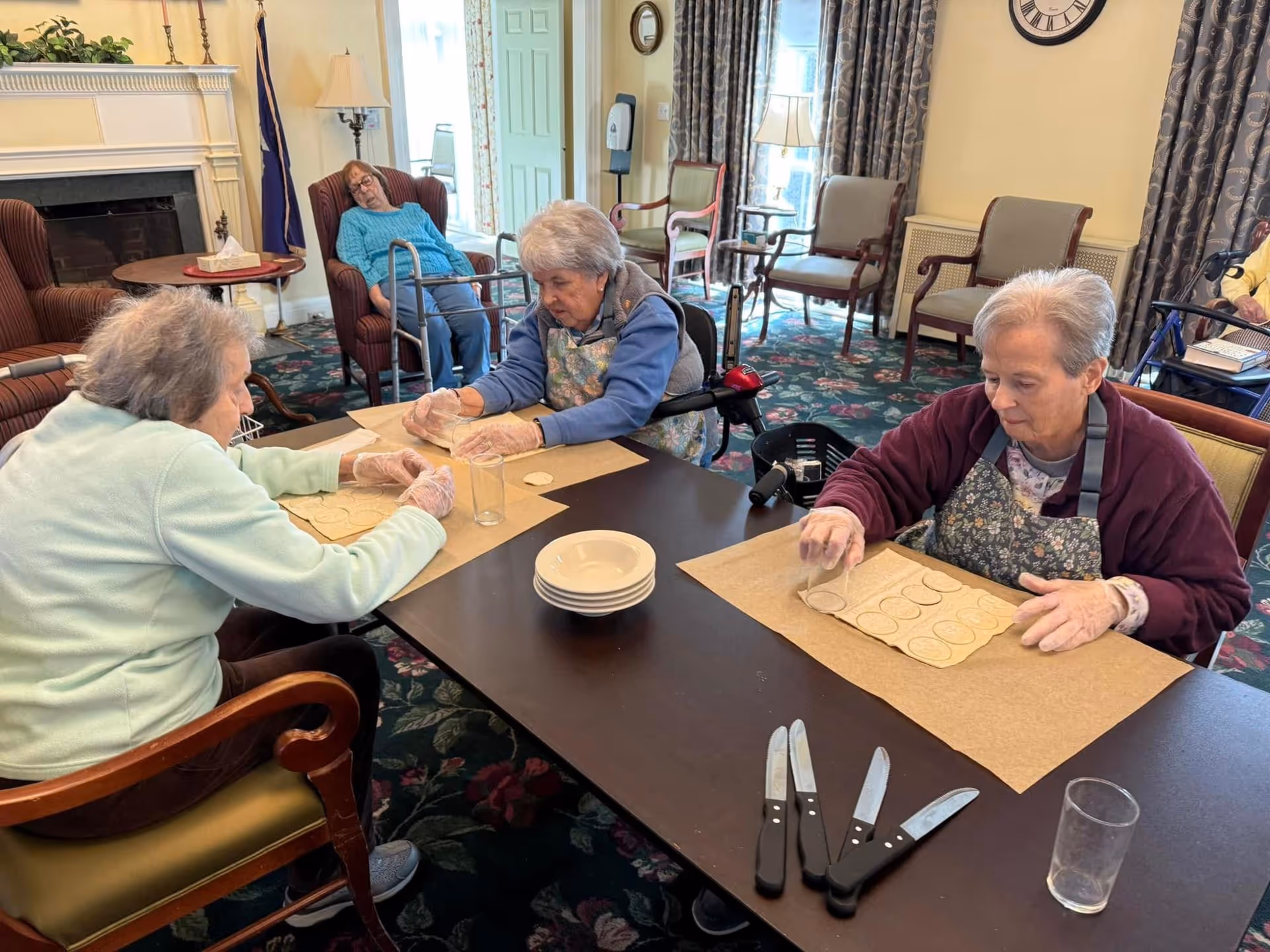 Three elderly women in a senior living common room sit at a table making circular cutouts while another resident rests in the background.
