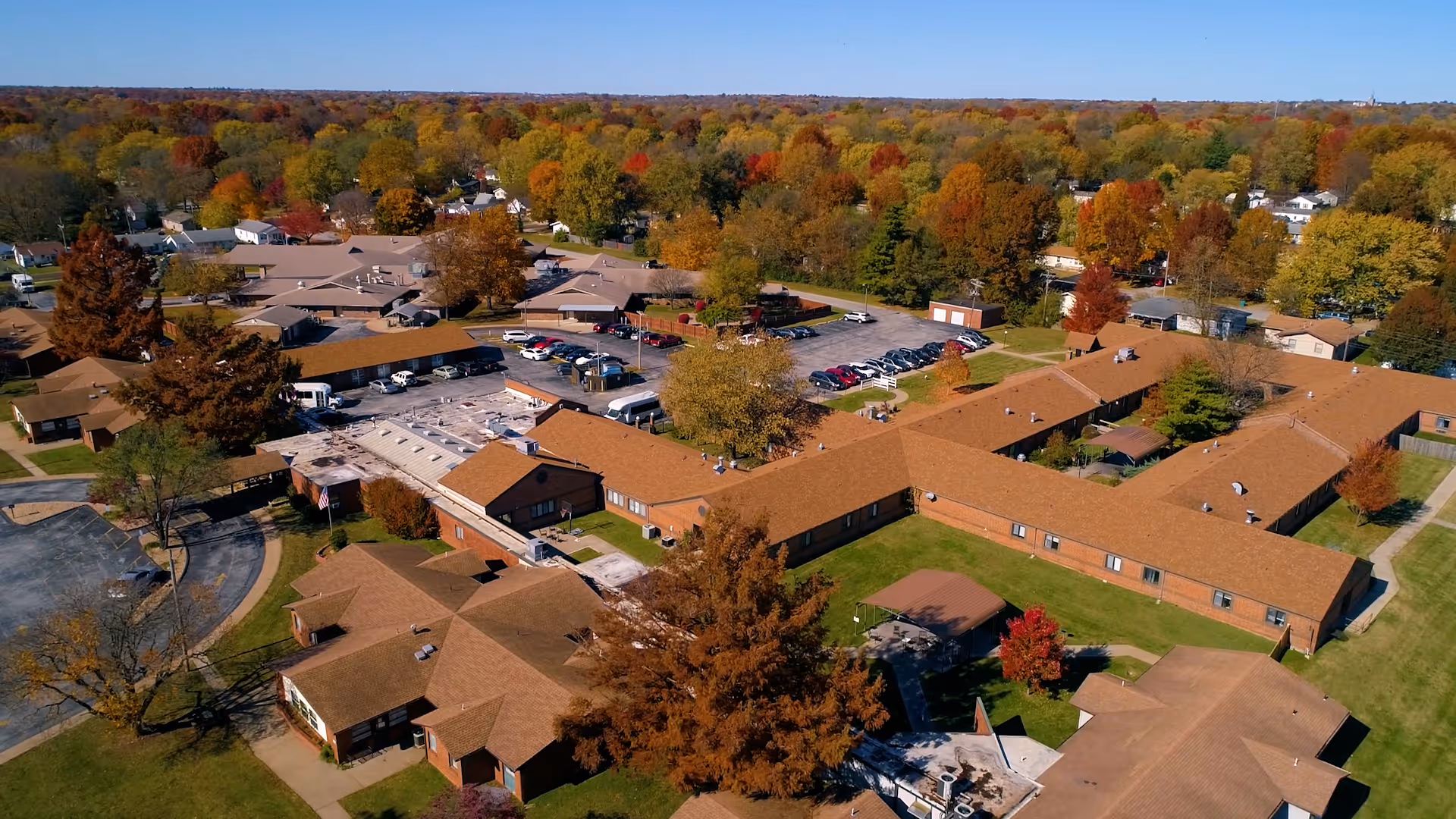 Aerial view of a senior care campus with connected single-story buildings, a parking lot, and surrounding autumn-colored trees.