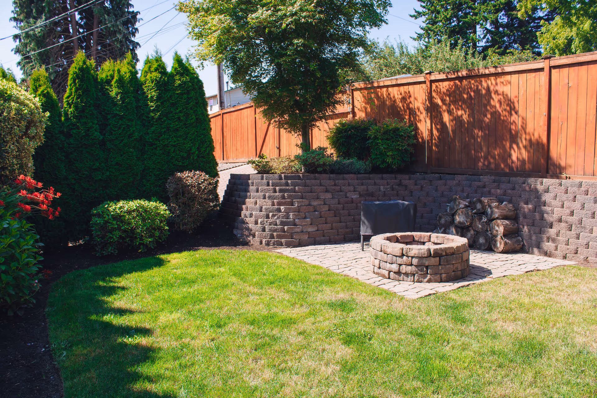 A sunny backyard with a circular brick fire pit, stacked firewood, a terraced retaining wall, wooden fence, and green lawn with shrubs.