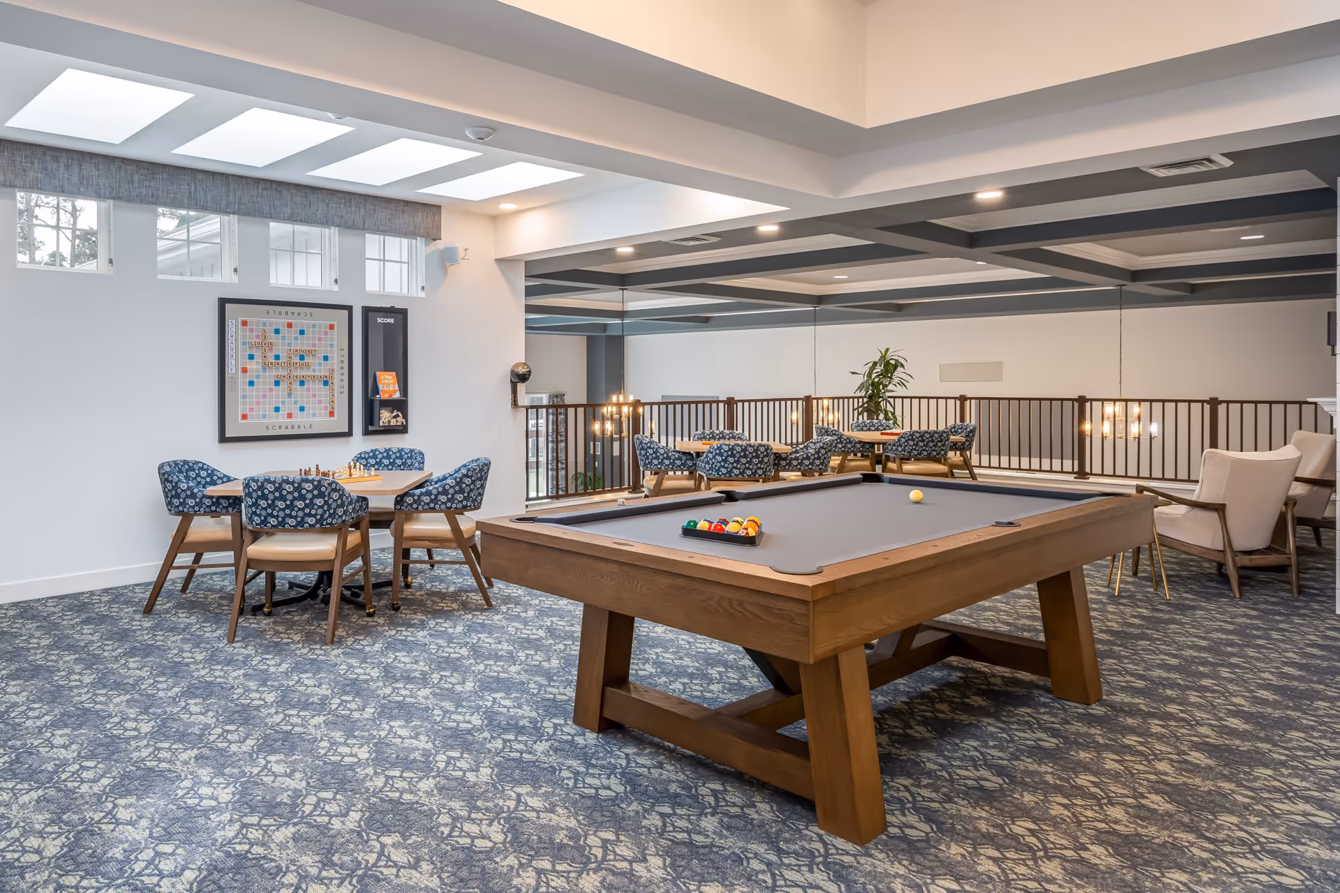 A senior living community game room featuring a pool table with balls set up, a chess and Scrabble game table with four blue patterned chairs, and additional seating areas with armchairs and tables. The room has skylights, a patterned carpet, and a railing overlooking a lower level.