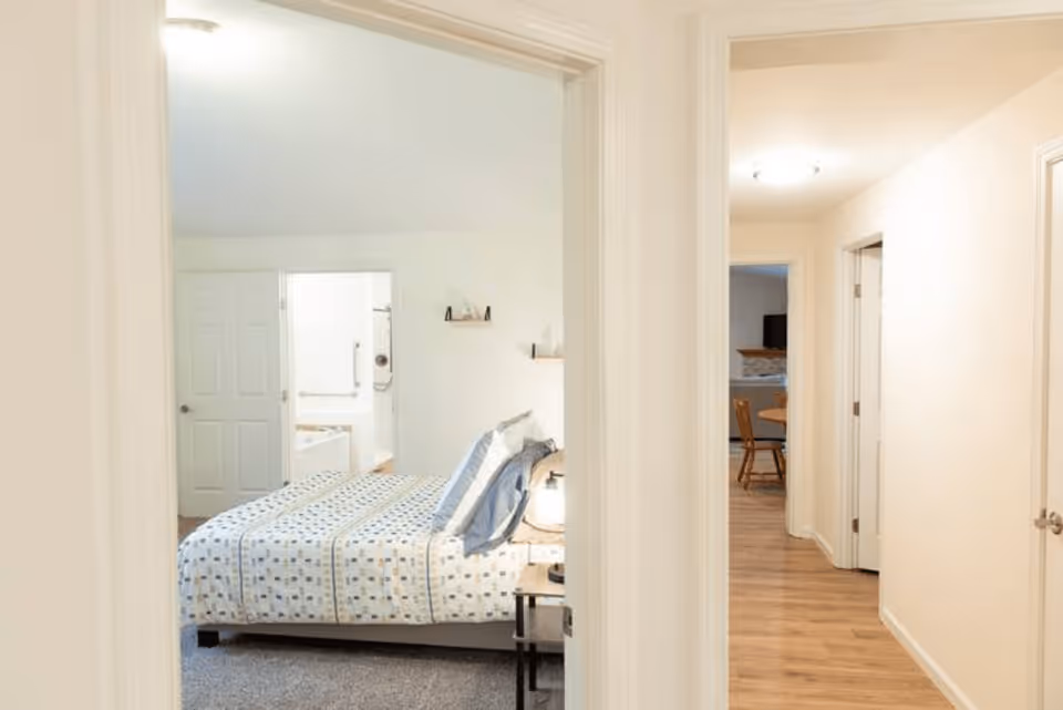 View through a doorway into a bedroom with a bed covered in a patterned bedspread and pillows. The bedroom has light-colored walls and carpeted floor. Beyond the bedroom, there is a bathroom with a bathtub and shower visible. To the right, a hallway with wooden flooring leads to another room with a round table and chairs.