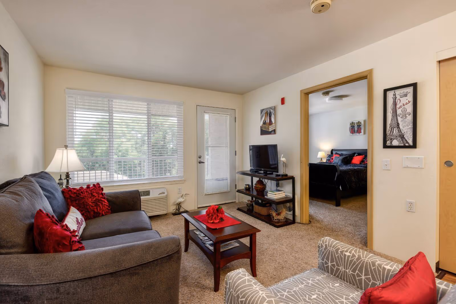 Living room with a gray sofa and armchair, coffee table, TV stand, and a doorway leading to a bedroom.