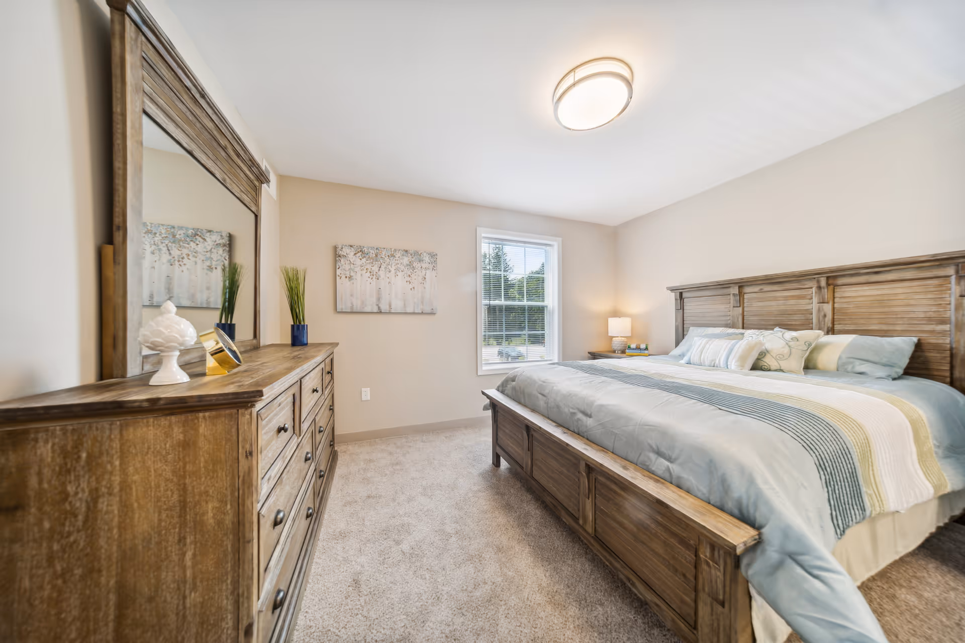 A bright bedroom with a large wooden bed featuring a blue and white striped comforter and multiple pillows. There is a wooden dresser with a large mirror on the left side, decorated with a small plant, a white decorative object, and a small clock. A window with blinds lets in natural light, and a bedside table with a lamp and books is visible on the right side. The walls are painted beige and the floor is carpeted.