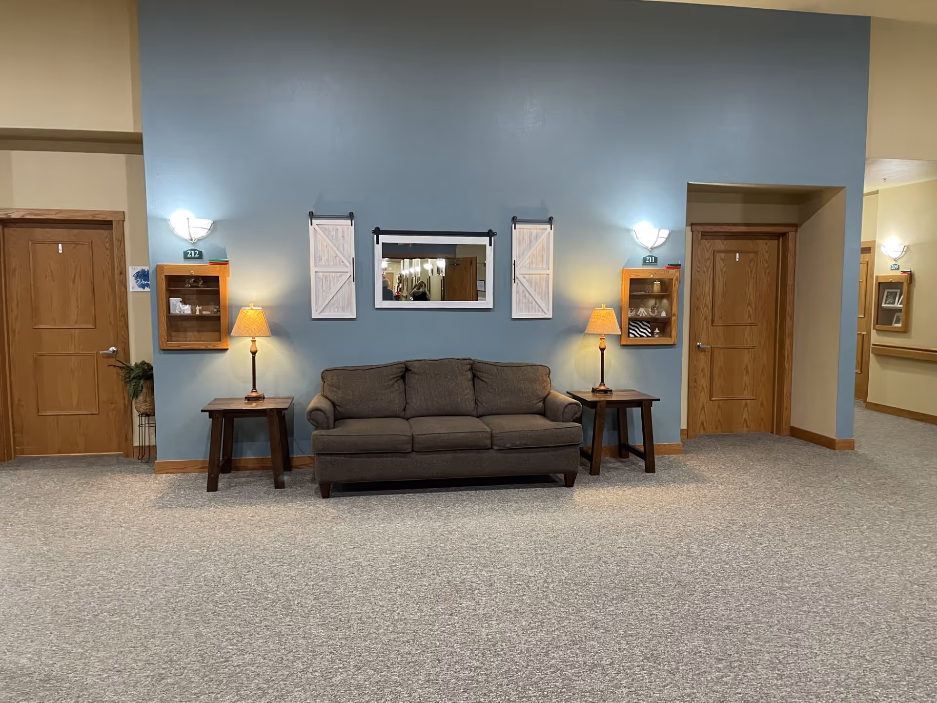Carpeted interior lounge with a brown sofa flanked by two side tables and lamps against a blue accent wall with a mirror and doors on each side.