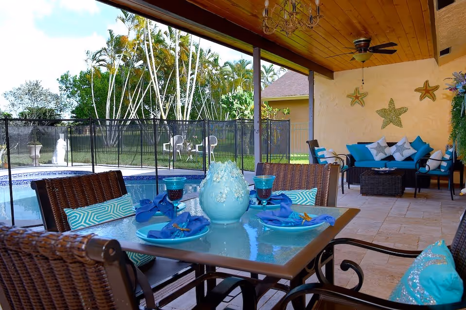 Covered patio dining and seating area with turquoise table settings overlooking a fenced pool and backyard.