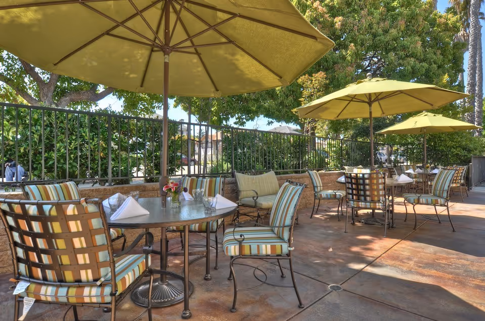 Outdoor patio area with several round tables, each surrounded by metal chairs with striped cushions. Large green umbrellas provide shade over the tables. There are trees and bushes along a fence in the background, and the area is paved with a concrete floor.