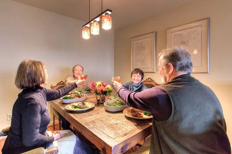 Four elderly people sitting around a wooden dining table in a warmly lit room, raising their glasses in a toast. The table is set with plates of food and a vase of flowers in the center. Two framed pictures hang on the wall behind them.
