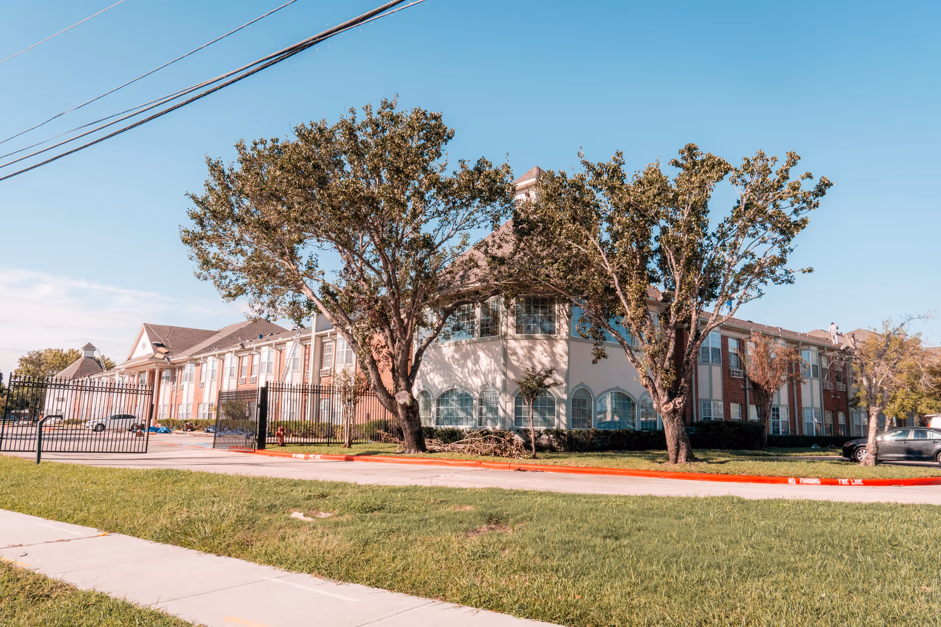 Front exterior of a two-story senior living building with trees, a gated driveway, and a grassy lawn.