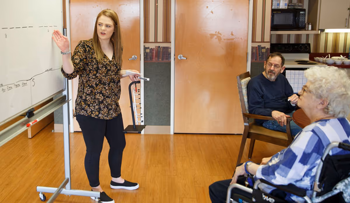 A woman stands near a whiteboard giving a presentation or lesson to two elderly individuals seated in chairs, one of whom is in a wheelchair, in a room with wooden floors and a kitchenette in the background.