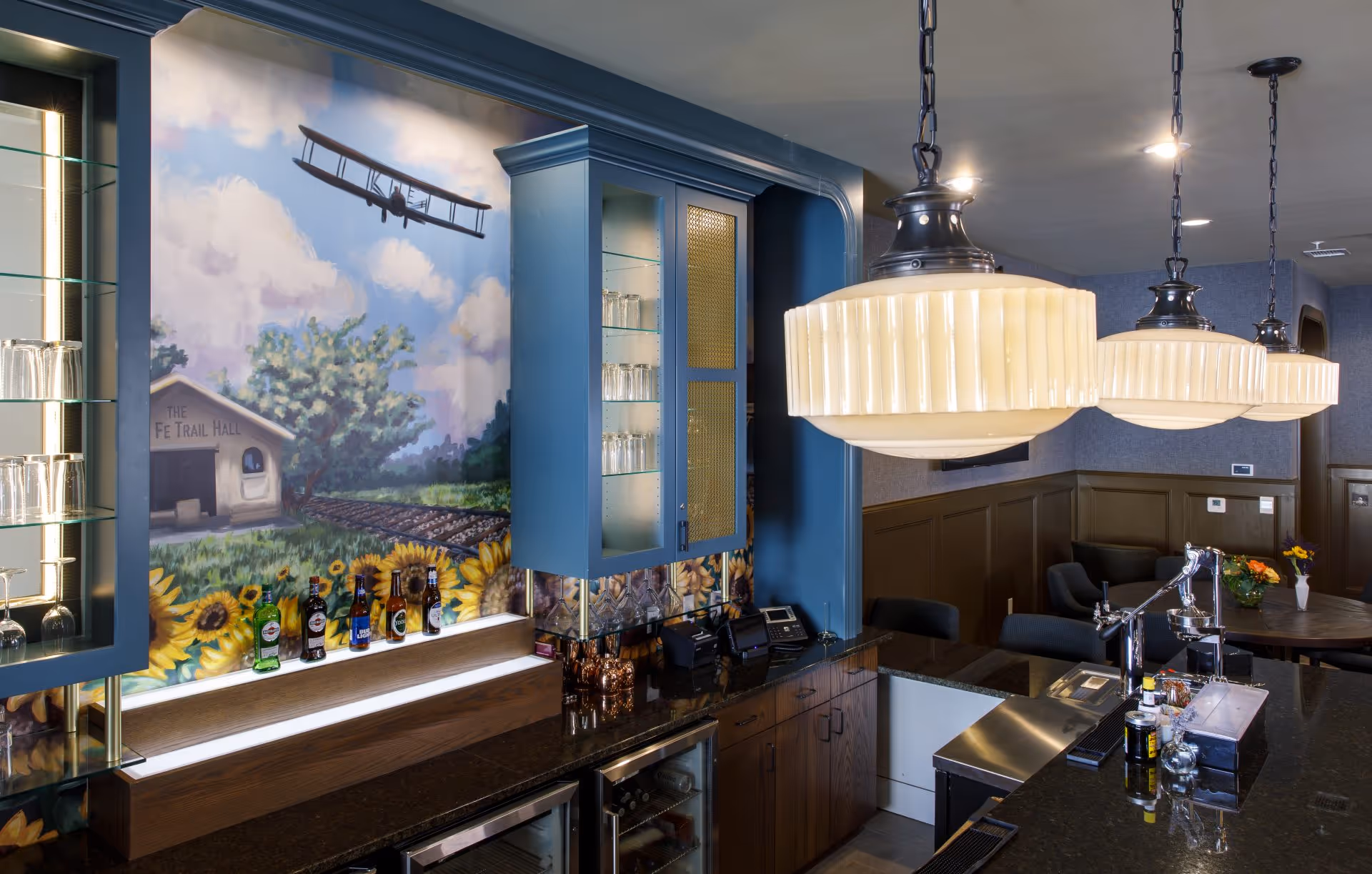 Interior view of a bar area with a mural on the wall depicting a vintage airplane flying over a building labeled 'The Fe Trail Hall' surrounded by sunflowers. The bar has glass cabinets with glasses, bottles of alcohol on the counter, and three large hanging pendant lights. In the background, there is a seating area with tables, chairs, and flower arrangements.