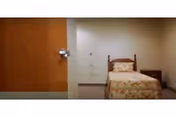Small institutional bedroom with a single bed with floral bedding, a nightstand, and a closed wooden door.