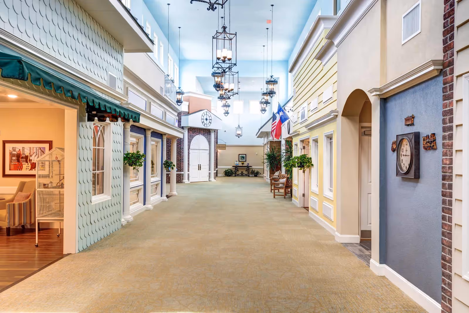 A bright, spacious indoor hallway designed to resemble a small town street with colorful building facades, hanging plants, benches, and multiple hanging lantern-style lights from a high ceiling with windows.