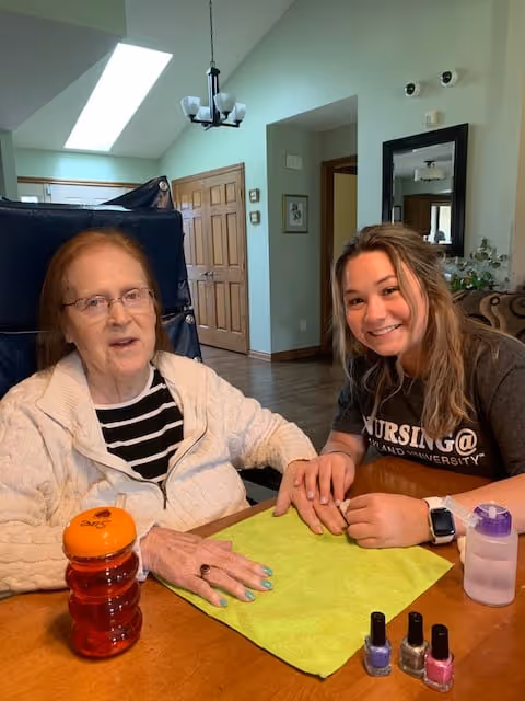 An elderly woman sitting at a table with a younger woman who is holding her hand and smiling. On the table are three bottles of nail polish, a green towel, a red drink bottle, and a water bottle. The setting appears to be a cozy, well-lit room with wooden doors and a chandelier in the background.