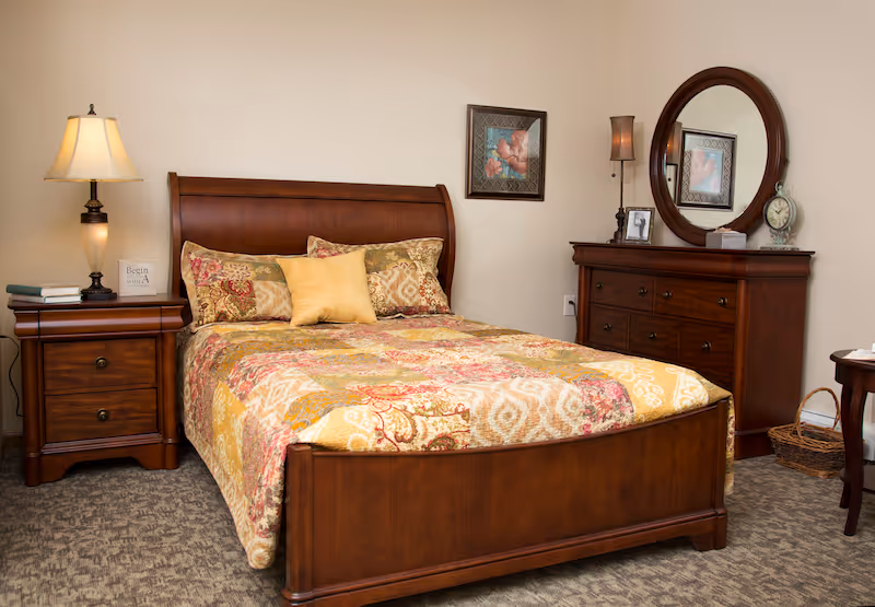 A warmly decorated bedroom featuring a wooden bed with patterned bedding, a nightstand with a lamp, and a dresser with a round mirror.