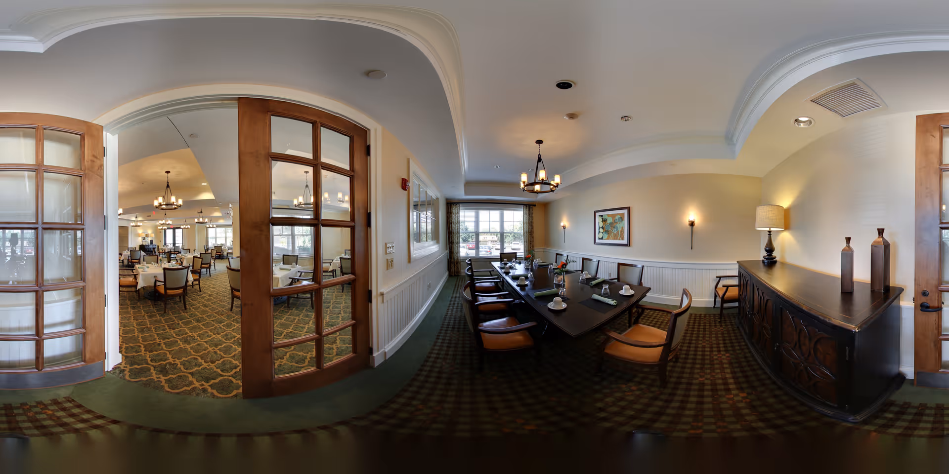 A spacious dining room with a long dark wooden table set for a meal, surrounded by cushioned chairs. The room features a patterned carpet, soft yellow walls with white wainscoting, a sideboard with decorative vases, and a large window letting in natural light. Adjacent to this room is another dining area visible through wooden framed glass double doors, with multiple tables and chairs arranged for dining.
