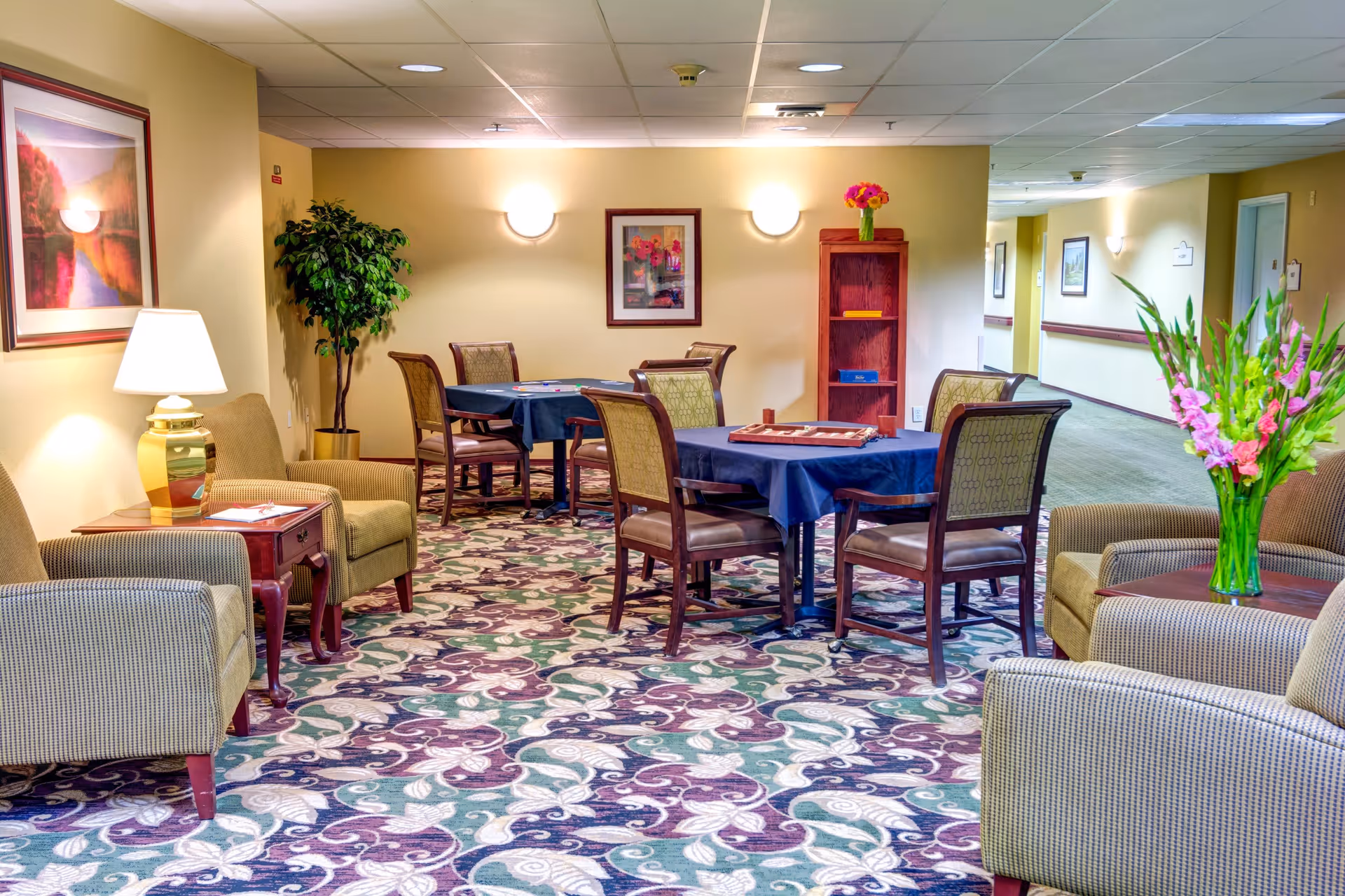 Brightly lit senior living common lounge with upholstered chairs, tables covered in blue cloths, patterned carpet, and floral arrangements.