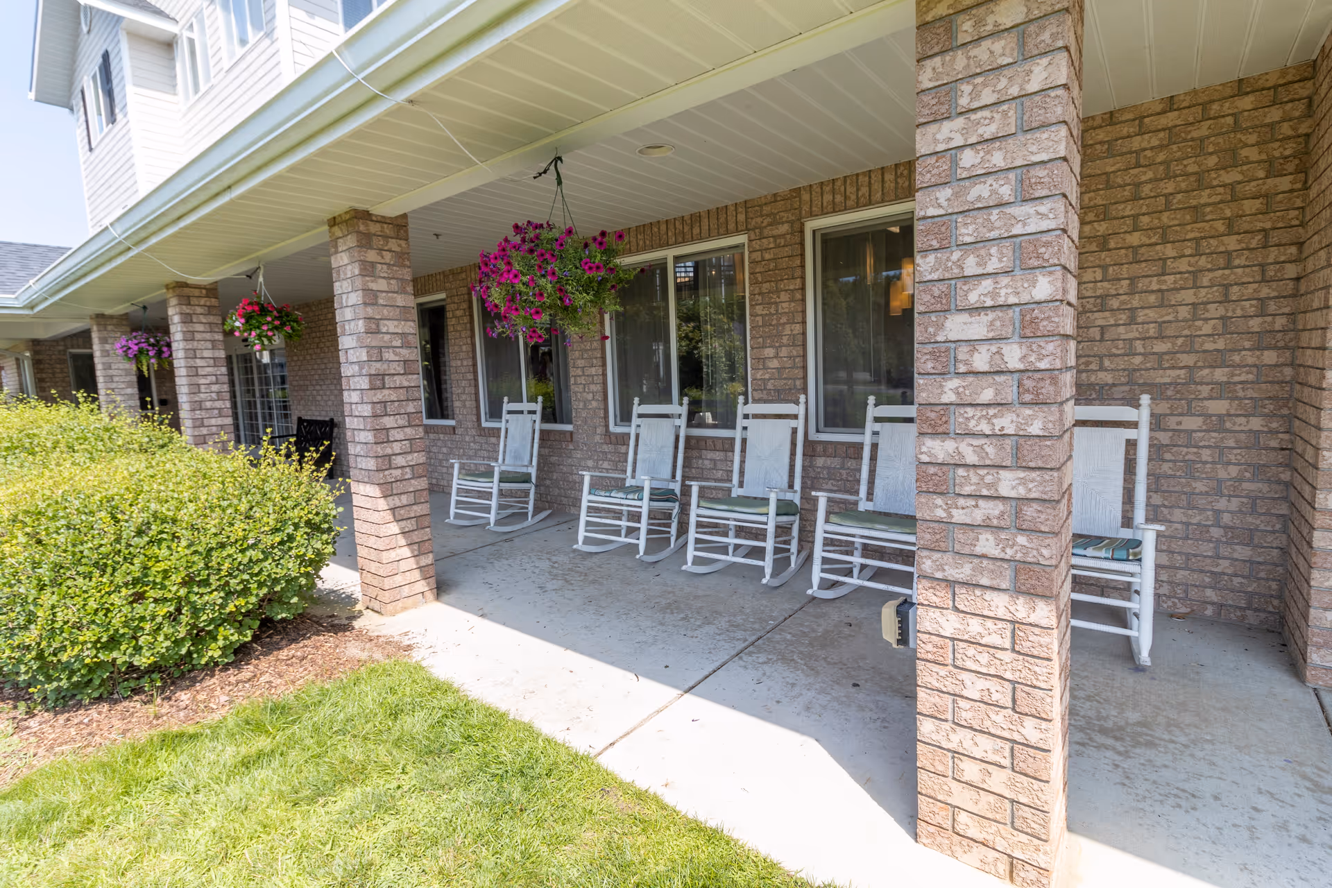 Covered outdoor patio area with several white rocking chairs lined up against a brick wall. Hanging flower baskets with purple and pink flowers are suspended from the ceiling. Green bushes and grass are visible in the foreground.