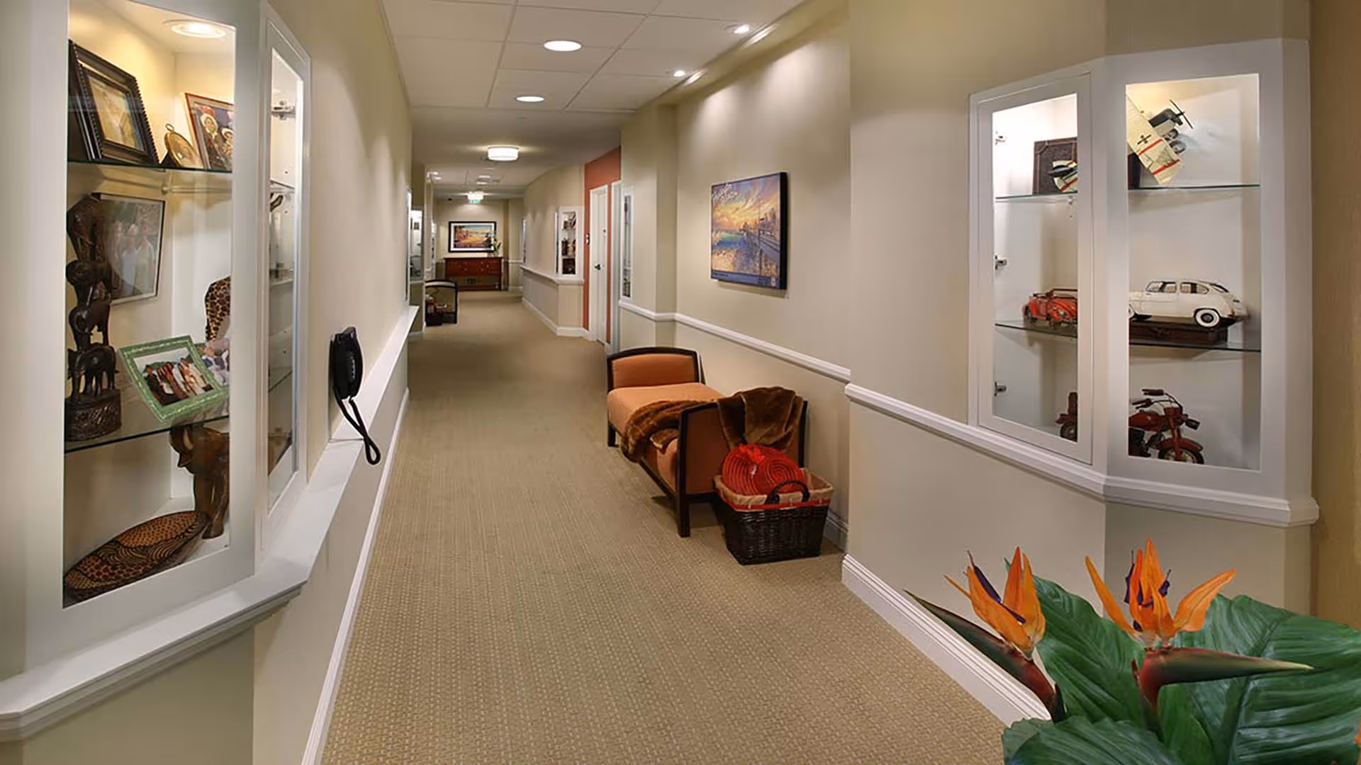 A well-lit hallway in a senior living facility with beige walls and carpeted floor. The hallway features display cabinets with decorative items such as model cars and framed photos. There is a bench with a blanket and a basket beside it, and a colorful painting on the wall. A plant with orange flowers is visible in the foreground.