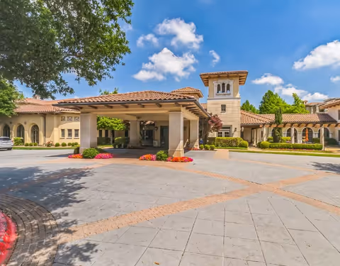 Exterior view of a senior living facility named Edgemere with a covered entrance, beige stucco walls, red tile roof, landscaped greenery, and a bright blue sky with scattered clouds.